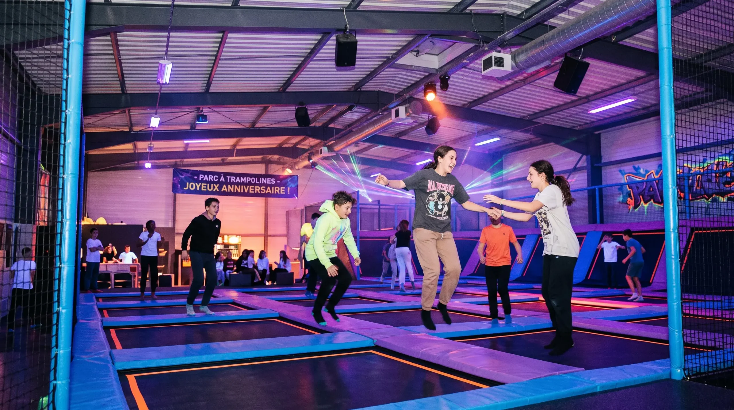Teenagers jumping on trampolines during a nighttime session in an indoor trampoline park with UV and neon lighting.