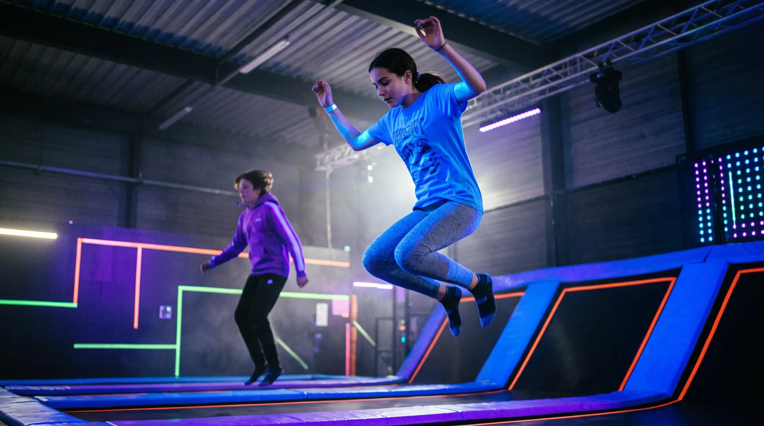 Two teenagers jumping on a trampoline under UV and neon lighting during a nighttime session in a trampoline park.